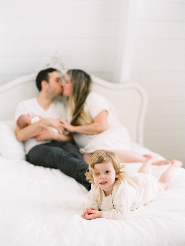 parents holding newborn baby on bed kissing with little girl playing