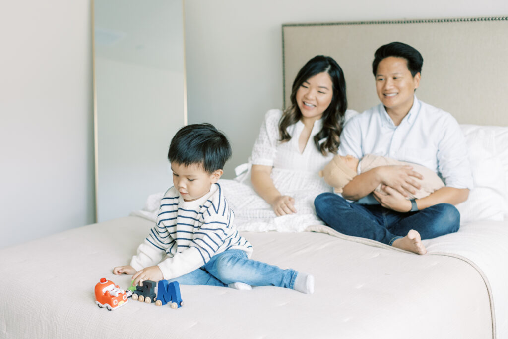 family photograph.  toddler son playing with toys, mother and father smiling at him holding newborn son
