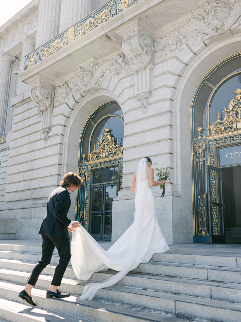 groom holding Brides wedding dress as they walk into san francisco city hall