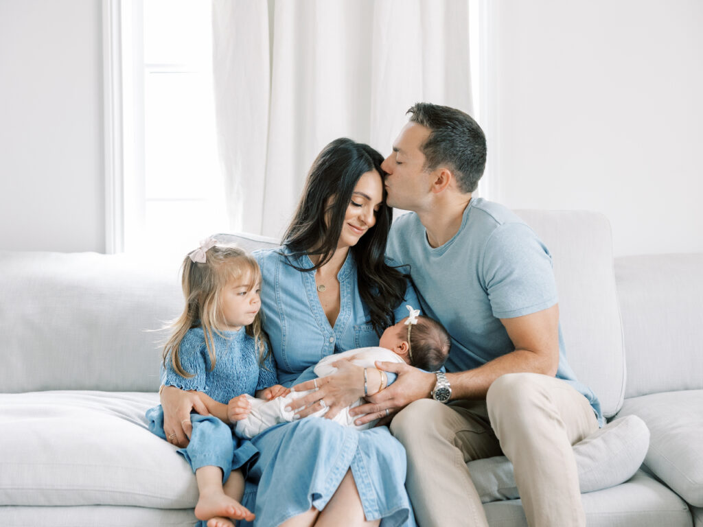 Bay Area Lifestyle Newborn Session with mother, father, toddler, and newborn.  father is kissing mother on the forehead while she holds the baby and sister looks over.